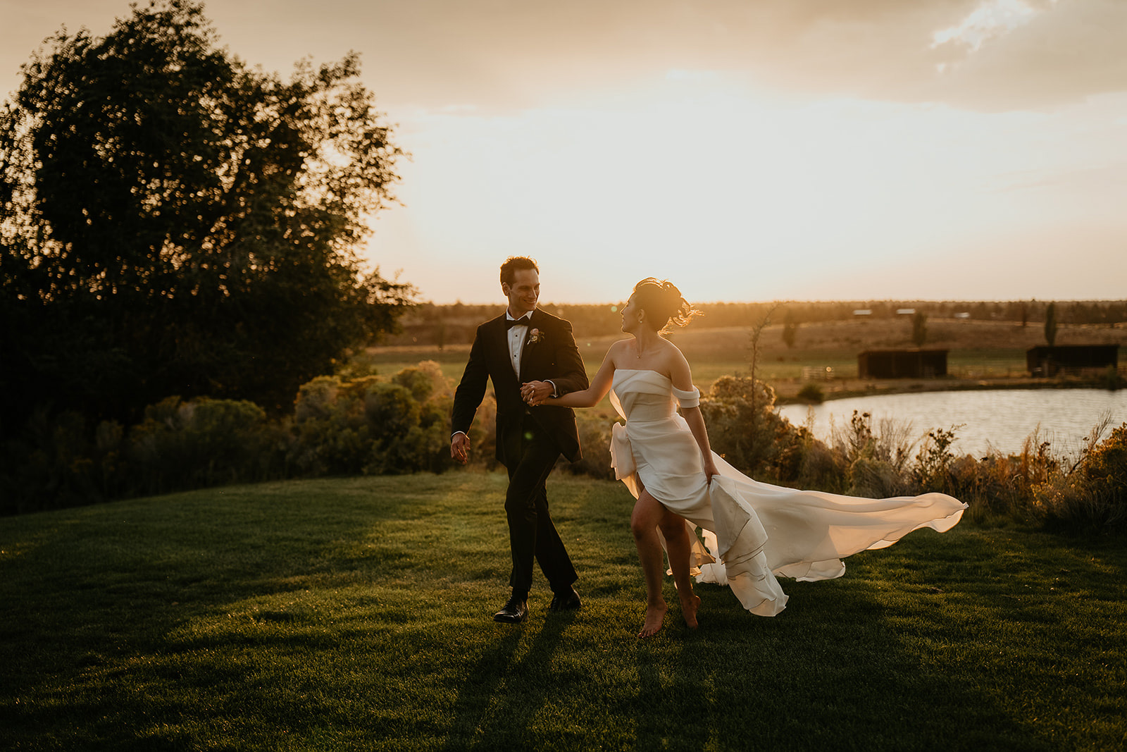 newlyweds walking on a grassy field with a pond in the background at sunset.