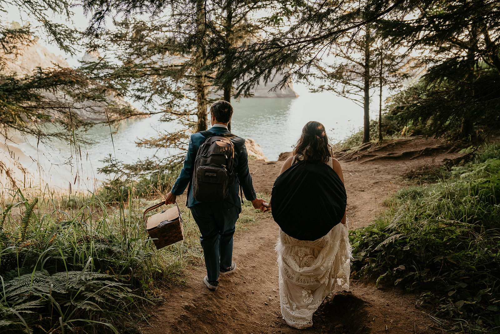 newlyweds holding hands as they walk along a trail by the coast. 
