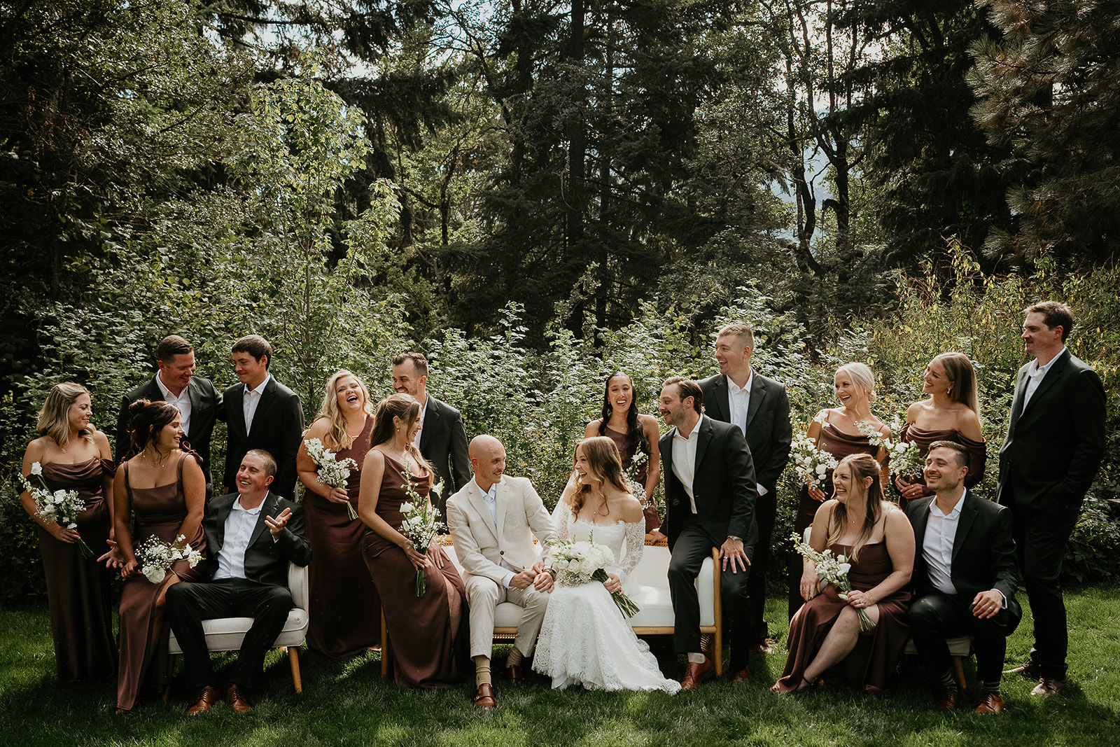 the bride, groom, and their wedding party on benches smiling with a forest in the background.