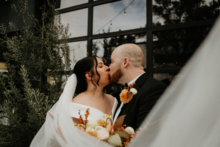 newlyweds kissing in front of a large window with her veil flowing in the wind during their Coopers Hall wedding.