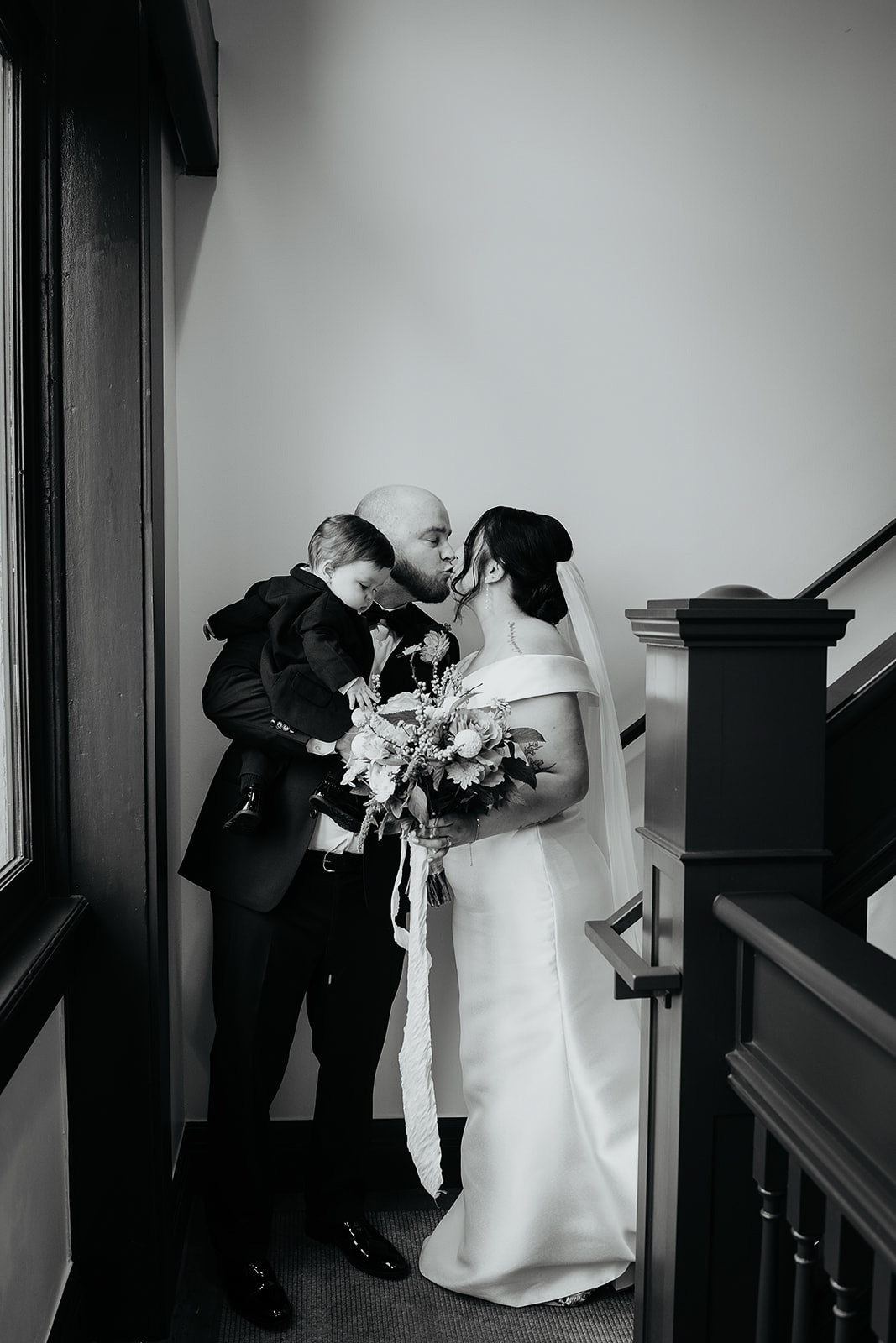 newlyweds kissing, the husband holding their son, in a staircase during their Coopers Hall wedding.