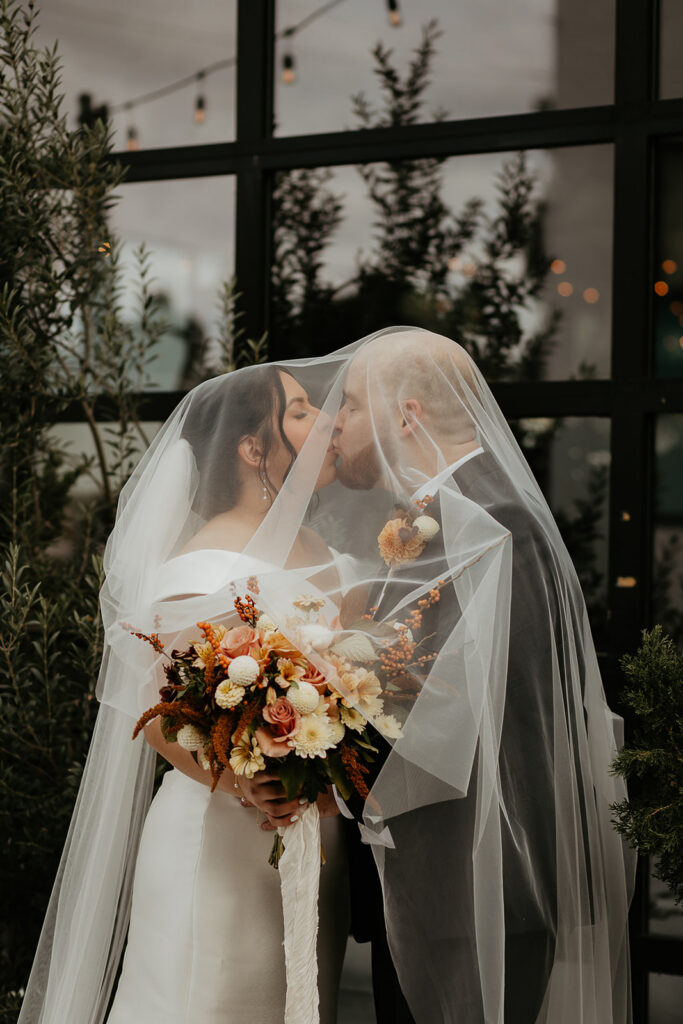 newlyweds kissing under the bride's veil in front of a large window during their Coopers Hall wedding.