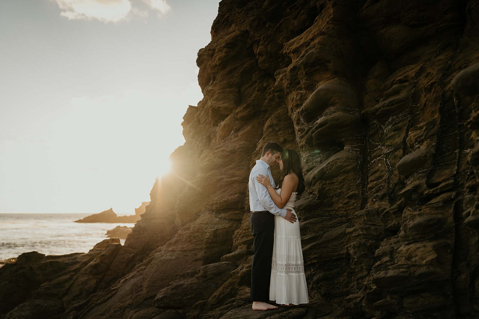 couple kissing as the sun sets behind a cliff on the beach.