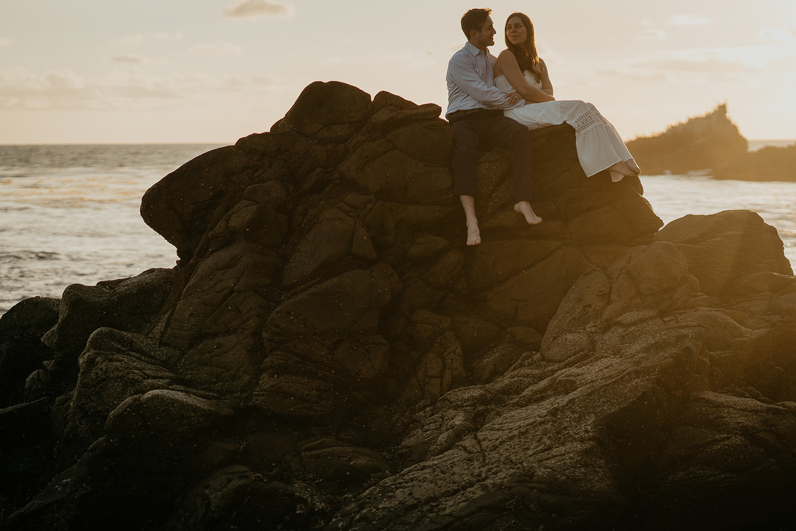 couple sitting on a rock outcropping with the ocean behind them.