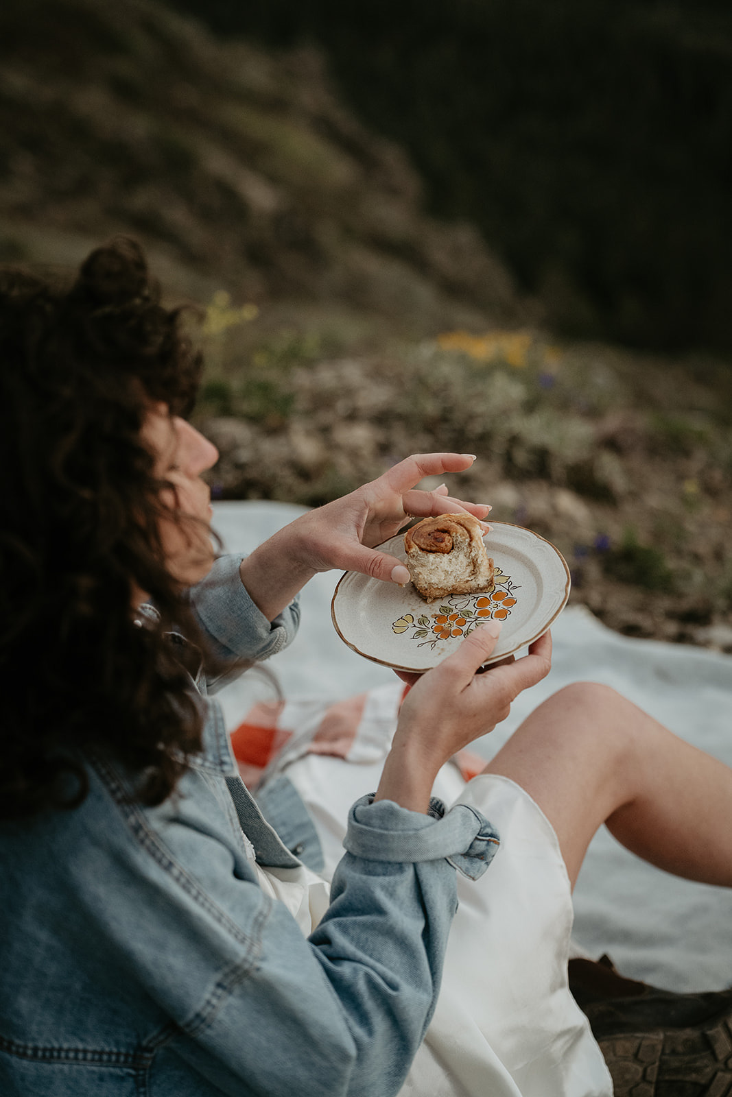 wife enjoying a cinnamon roll while sitting on a picnic blanket in the mountains during their wedding anniversary photoshoot.