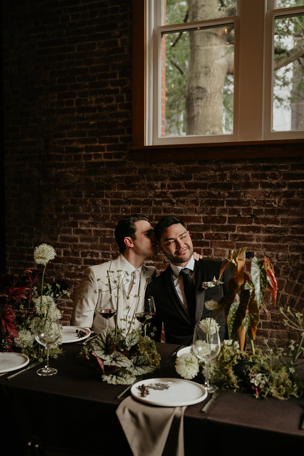 Newlyweds kissing at a decorated wedding table in front of a brick wall.