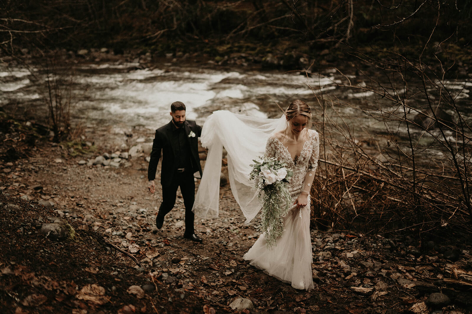 Groom holding bride's veil while walking along the riverbank