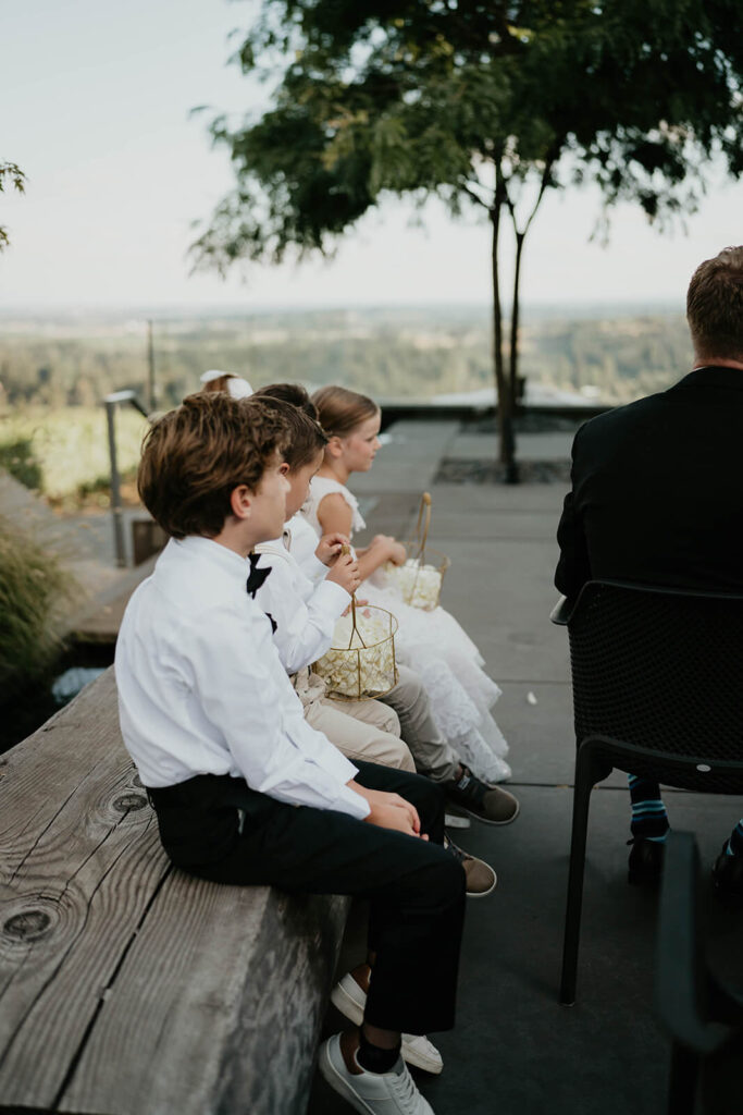 Flower girls and ring bearers sitting on wood bench during outdoor wedding ceremony