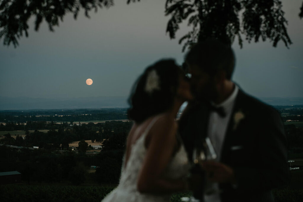 Bride and groom holding wine glasses and kissing during blue hour
