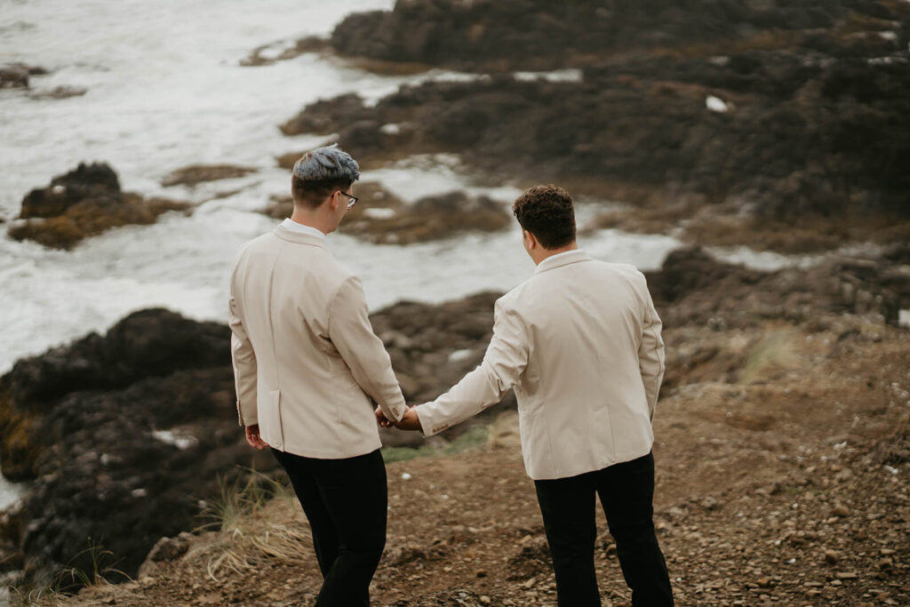 Two grooms holding hands and hiking down their elopement ceremony site on the Oregon Coast