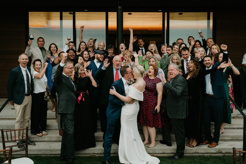 Bride and groom kiss while friends and family cheer during wedding portraits