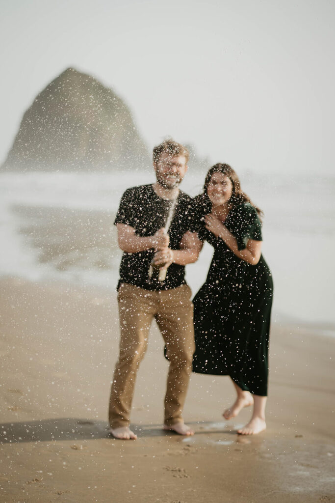 Couple laughing and popping champagne on Cannon Beach during their beach engagement photos