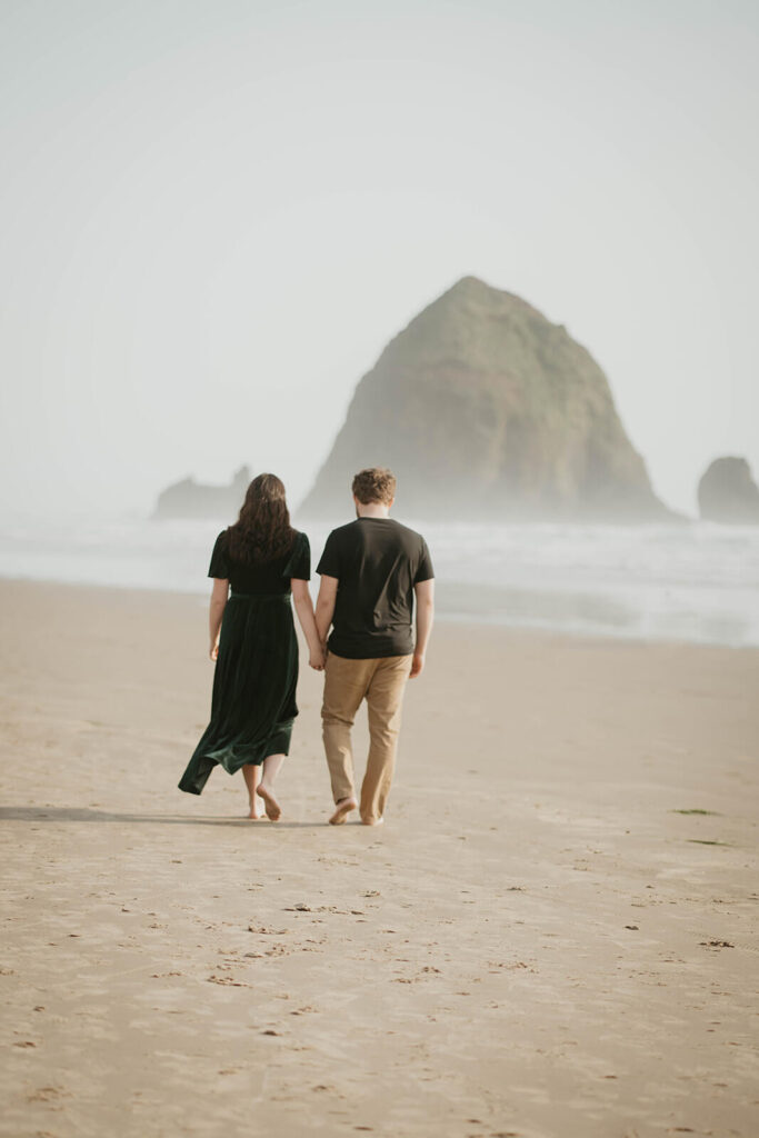 Couple holding hands, walking across Cannon Beach during their beach engagement photos