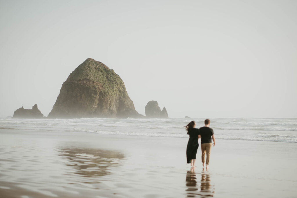 Couple holding hands walking across Cannon Beach in Oregon for their beach engagement photos