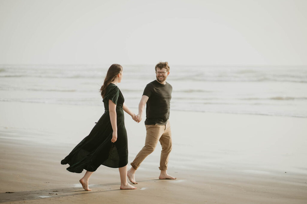Couple holding hands, walking across Cannon Beach during their beach engagement photos