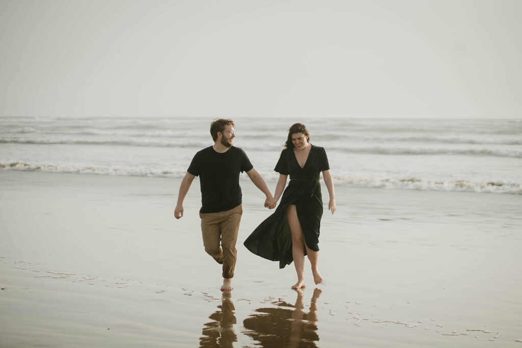 Couple holding hands, running across Cannon Beach during their beach engagement photos