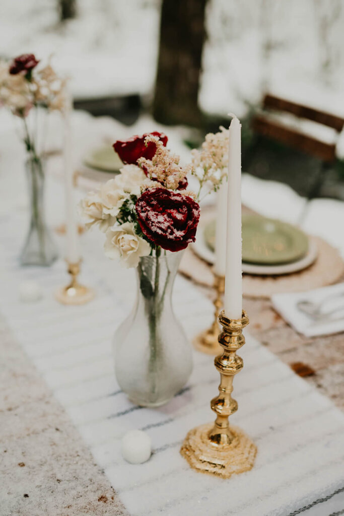 White and burgundy florals in a bud vase sitting next to white candles in gold candlestick holders at winter wonderland cabin elopement