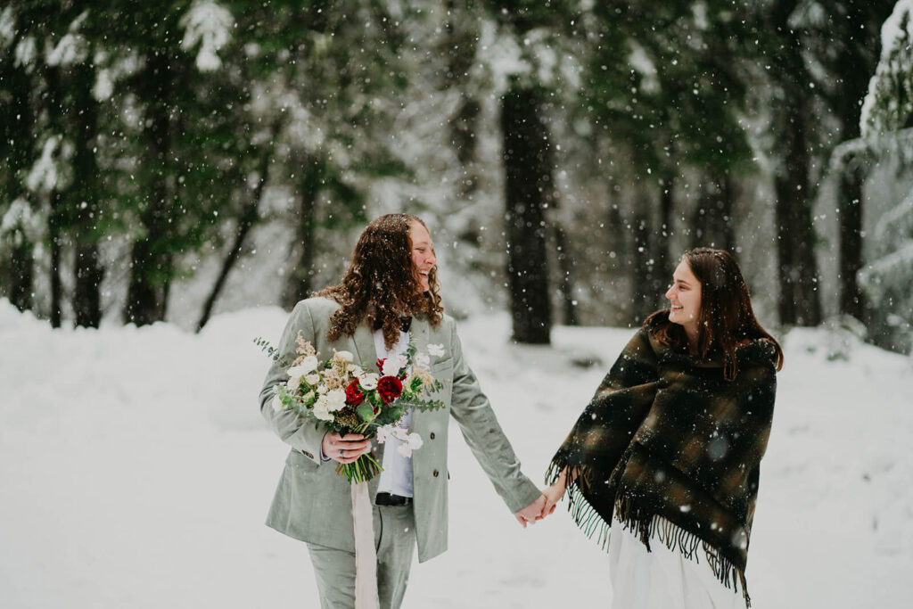 Two brides holding hands, walking through the snow at winder wonderland cabin elopement in Oregon