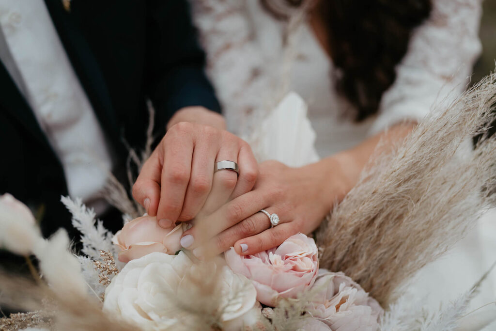 Bride and groom wearing wedding rings resting their hands on neutral floral wedding bouquet