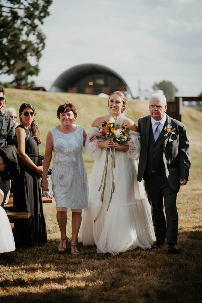 Bride walks down outdoor wedding aisle with her parents