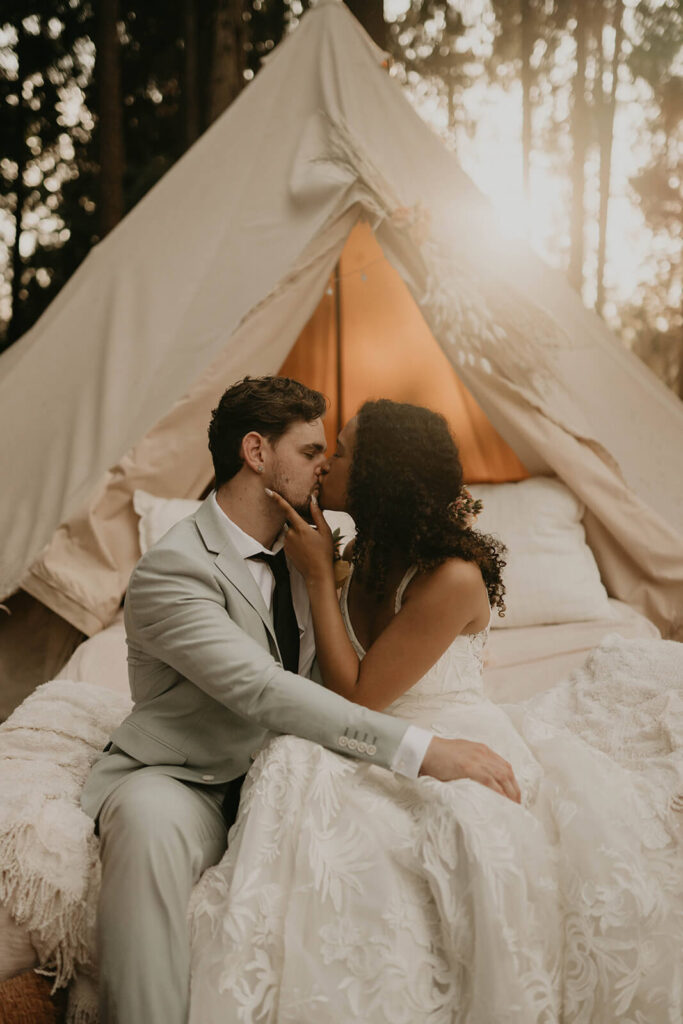 Bride and groom kiss while sitting on an outdoor bed in the woods surrounded by orange pillows and a white linen tent