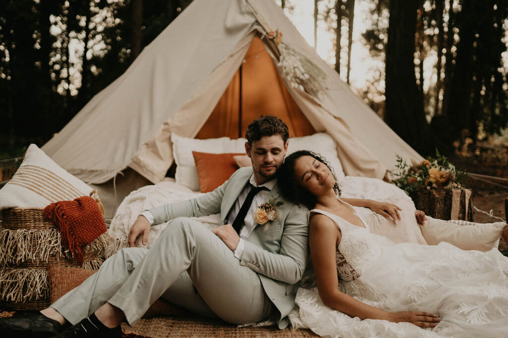 Bride and groom relax on the ground in front of a white bed in the woods surrounded by orange pillows and a beige linen tent