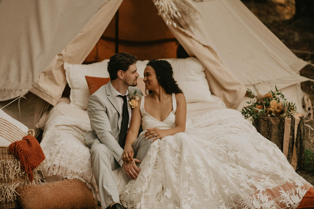 Bride and groom sit on an outdoor bed in the woods surrounded by orange pillows and a white linen tent