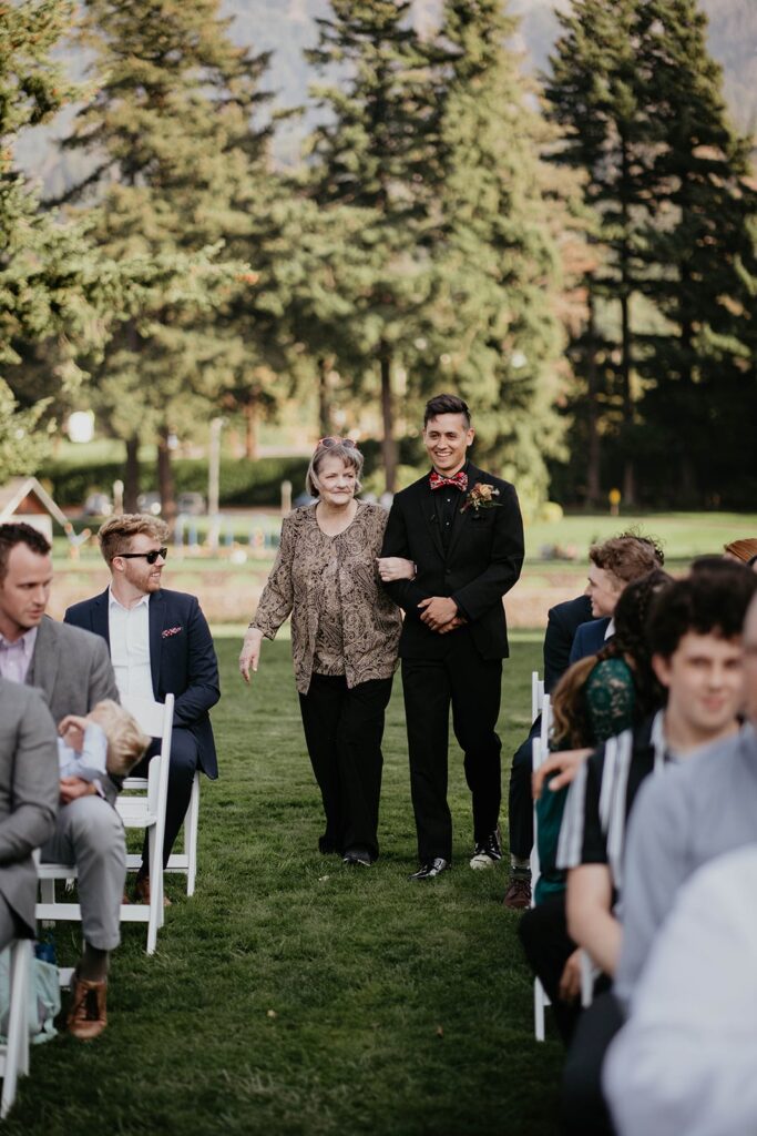 Groom entering outdoor wedding ceremony with mother on his arm