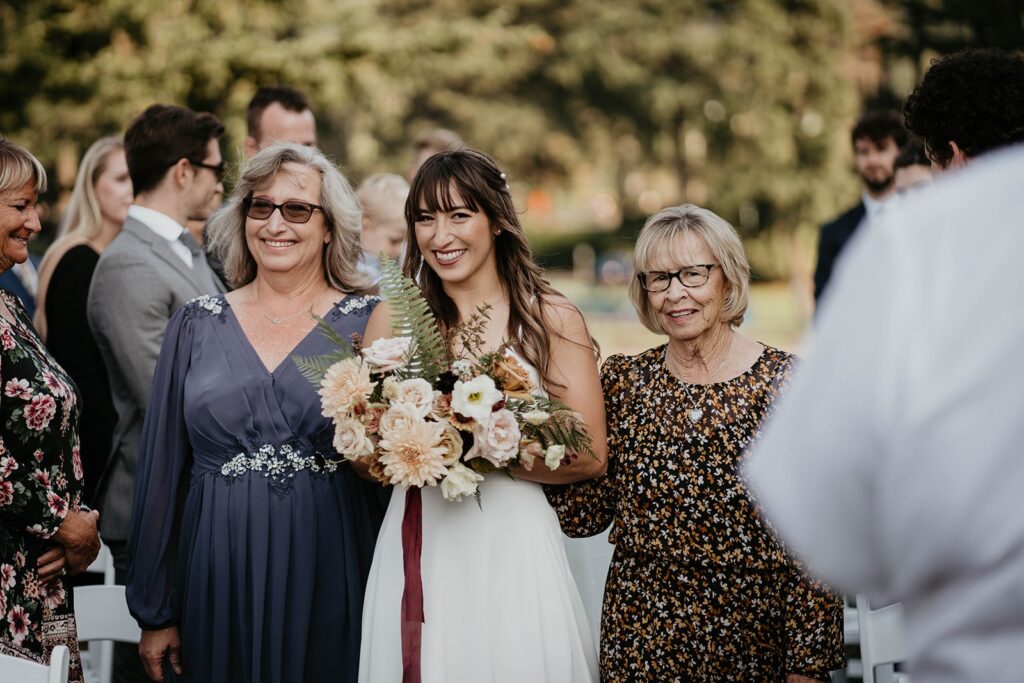 Groom smiling as she enters the outdoor wedding ceremony with her mother and grandmother