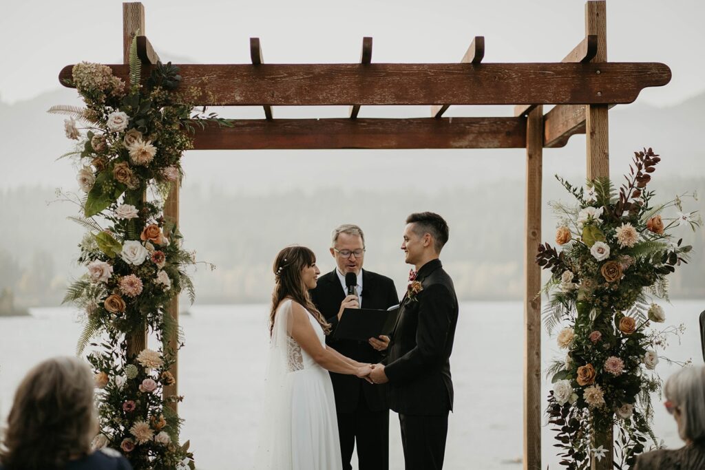 Bride and groom standing under a wood arch decorated with orange, pink, and green florals during their Thunder Island wedding