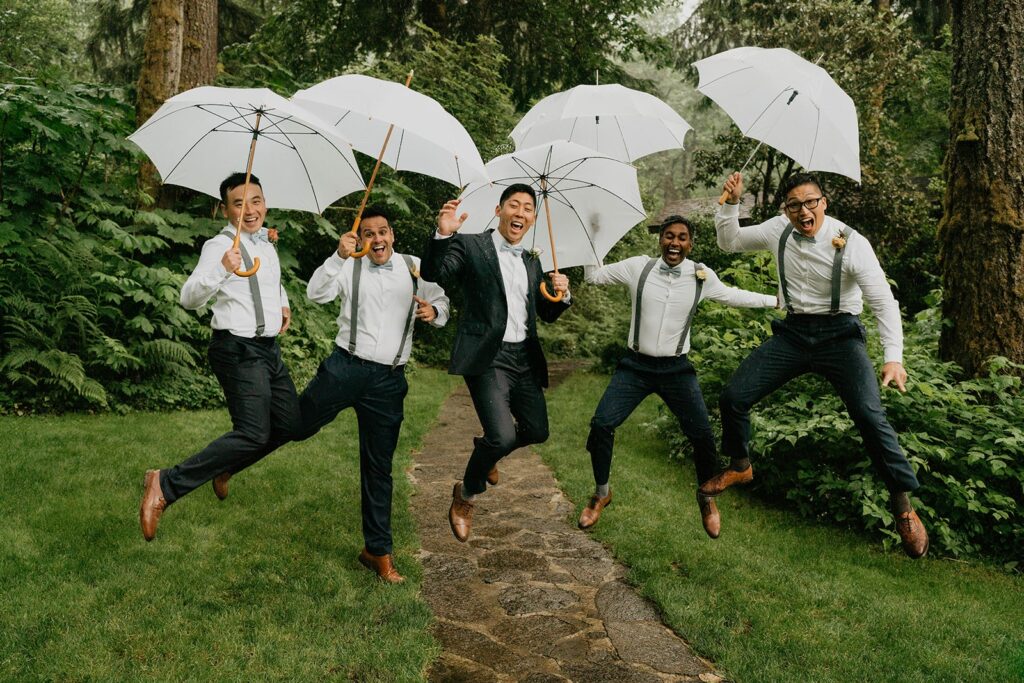 Groom and groomsmen jump while holding white umbrellas at Bridal Veil Lakes wedding