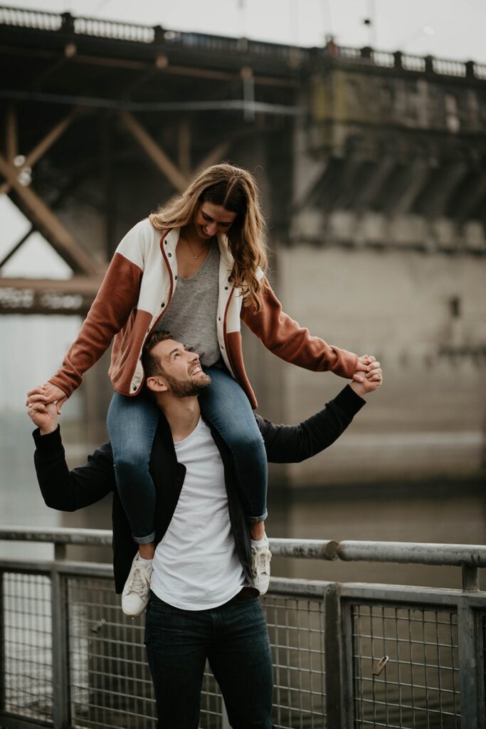 Woman riding on man's shoulders during Portland engagement photo session by the river