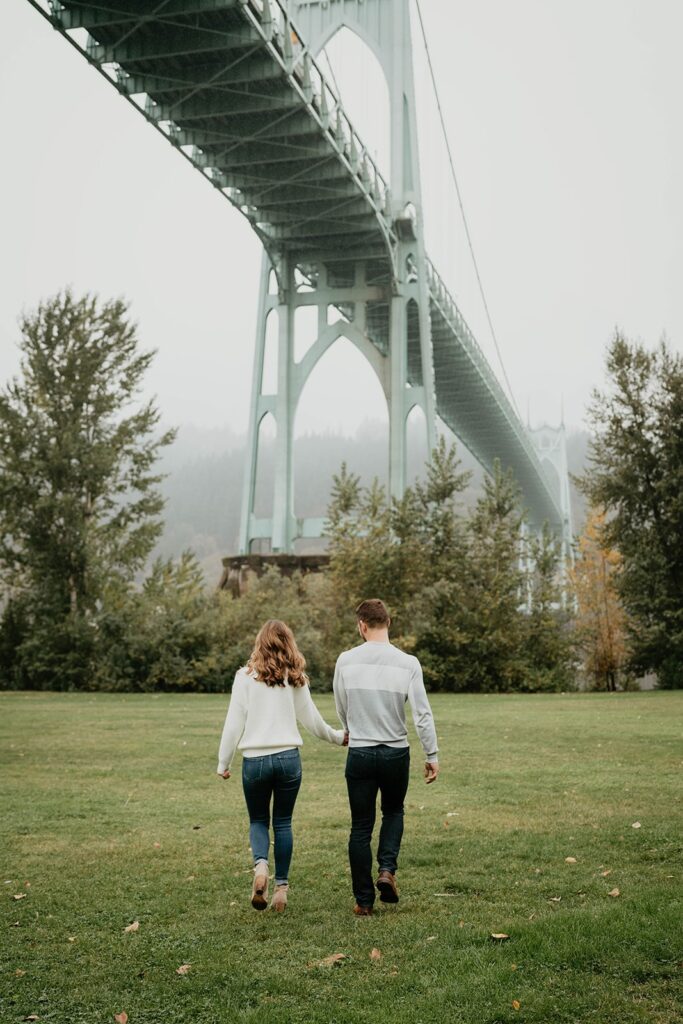 Couple holding hands while walking across the grass during Cathedral Park, Oregon engagement session