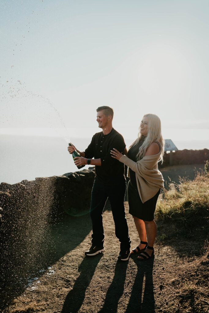 Couple popping a bottle of champagne at Cape Perpetua viewpoint for their Oregon Coast engagement photos