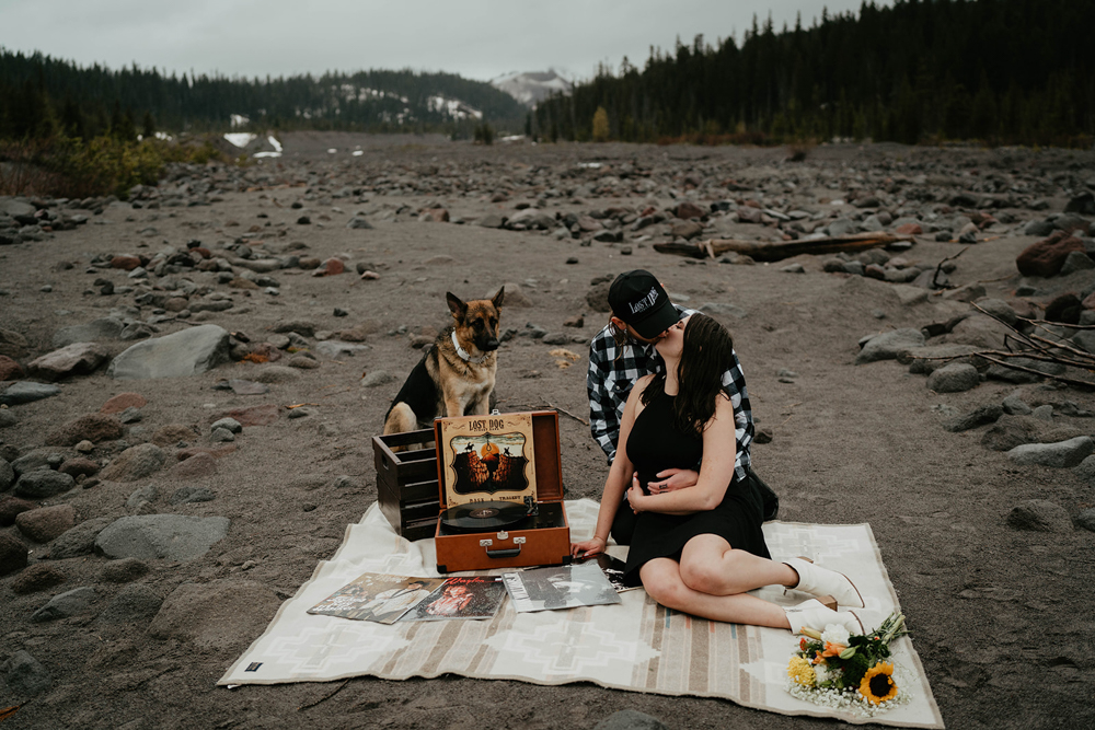 A couple kissing, sitting on a picnic blanket with records and their dog, and Mt. Hood in the background at White River West Sno-Park.