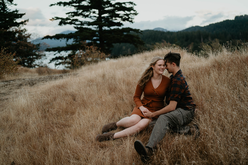 A couple cuddling in a field with a lake in the background at Government Cove. 