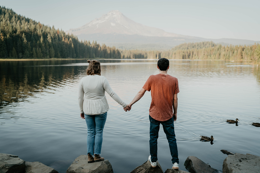 A couple holding hands while standing on a pathway of rocks, with Trillium Lake and Mt. Hood in the background. 