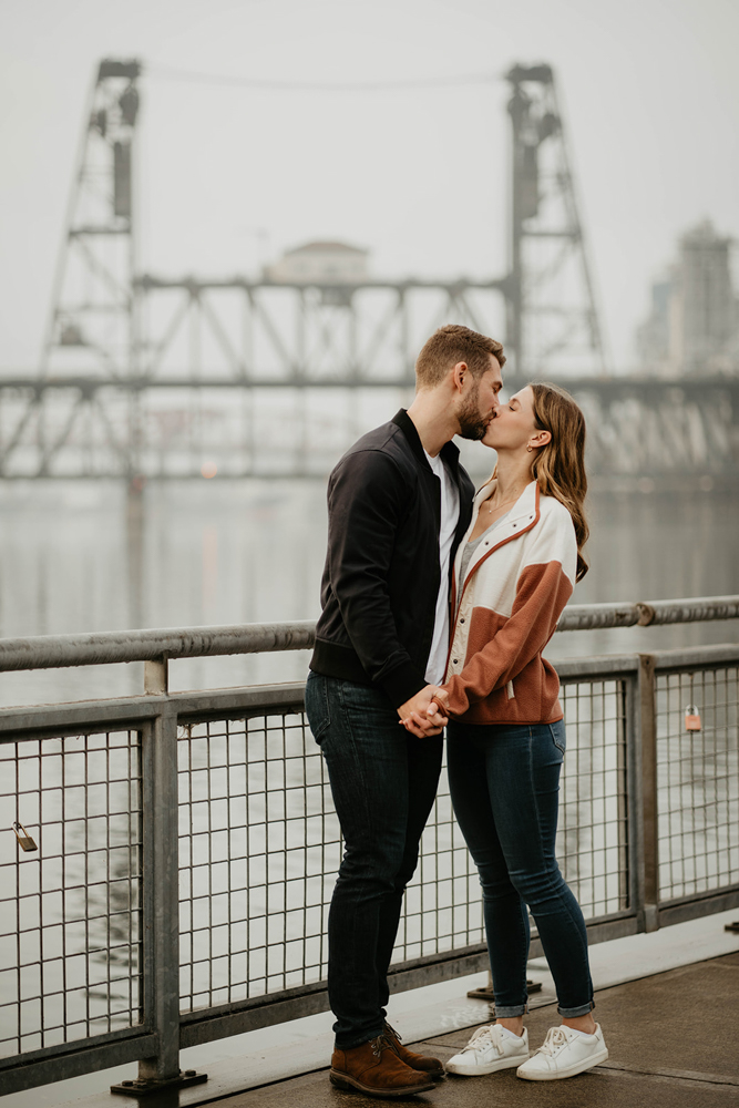 A couple kissing in front of a steal bridge along the Willamette River Waterfront in Portland. 