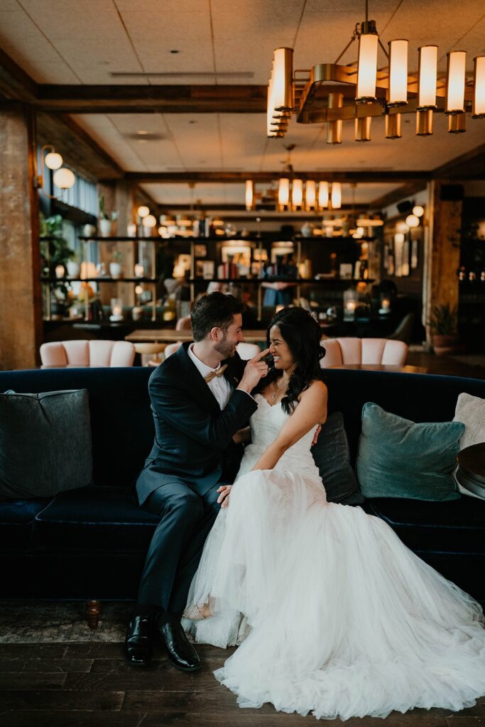 Bride and groom laugh while sitting on a blue velvet couch at Blockhouse PDX for their wedding portraits