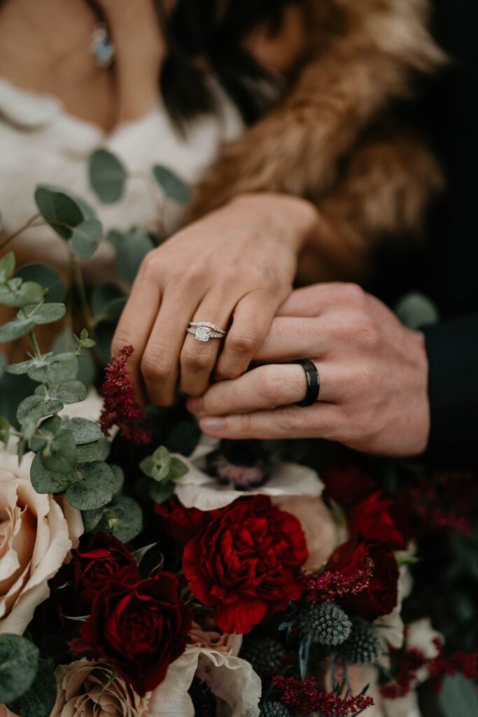 Bride and groom rest hands with diamond and black wedding bands on top of red, white, and green flower bouquet.