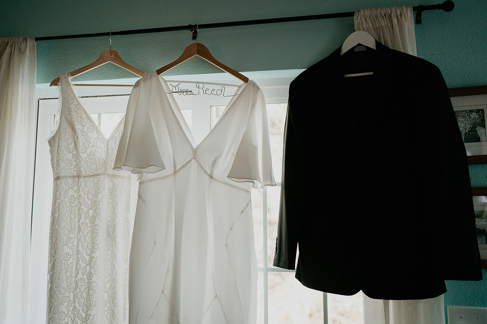 A wedding dress and jacket hanging with a teal wall and window in the background. 