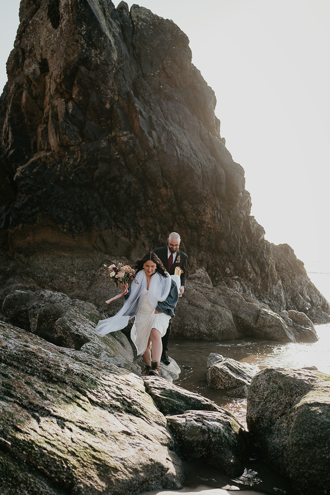 The bride holding flowers leading the groom up onto sea rocks at Cannon Beach. 