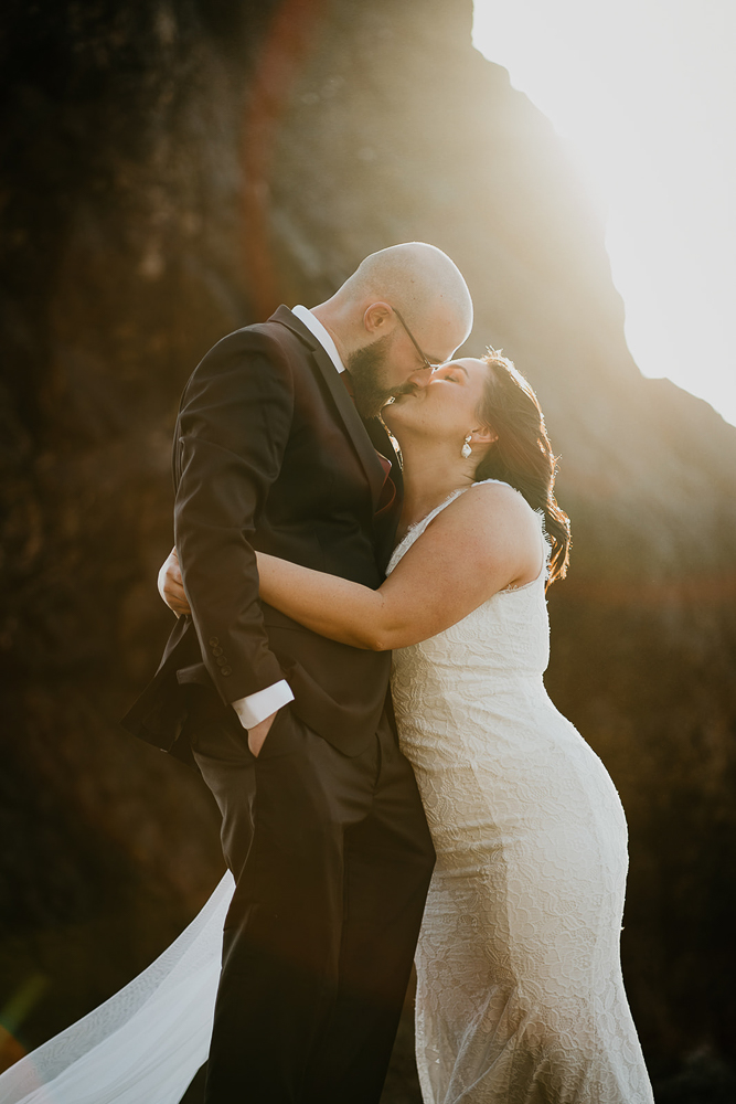 The bride and groom kissing with the sun shining in the background while standing on a sea stack at Cannon Beach. 