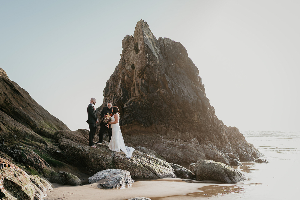 The bride, groom, and officiant standing on a sea stack at Cannon Beach. 