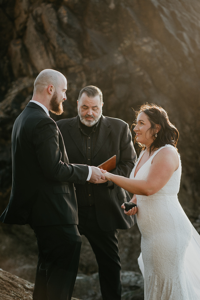 The bride and groom exchanging rings with the officiant overseeing while standing on a sea stack at Cannon Beach. 