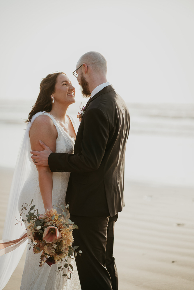 The newlyweds admiring each other, with the bride holding a bouquet of flowers while both are standing in the sand at Cannon Beach. 