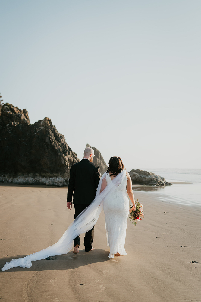 The newlyweds walking towards a sea stack at Cannon Beach, with the bride's veil flowing in the wind. 