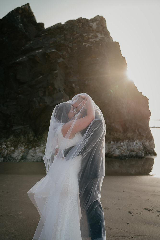 The bride and groom hugging, with the bride's veil covering both of them at Cannon Beach. 