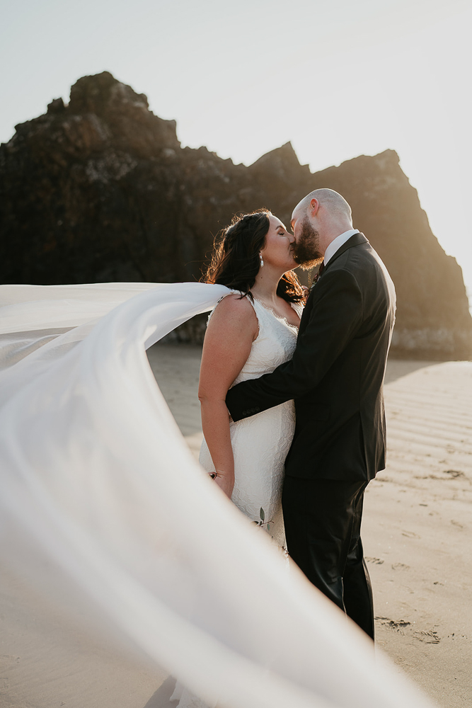 The newlyweds kissing while the bride's veil flows in the wind at Cannon Beach. 