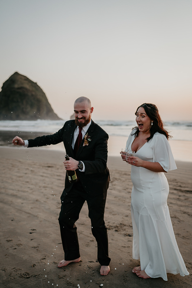The groom popping open a beer bottle while the bride looks on astonishingly at Cannon Beach. 